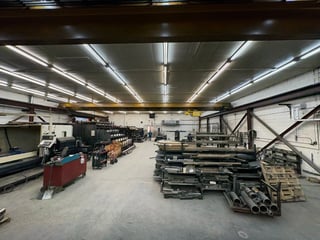 Industrial warehouse interior with machinery, metal materials, and equipment organized in a spacious manufacturing facility with bright overhead lighting.