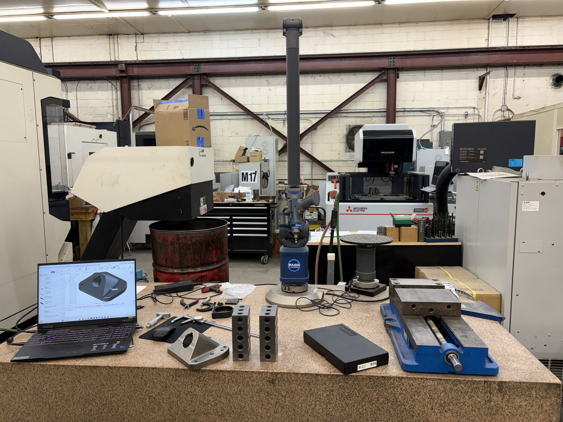 An industrial workspace with 3D printers and manufacturing equipment, including a laptop displaying a model and various tools and parts on the workbench