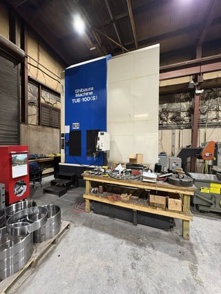 Industrial workshop with large blue and white CNC machine, wooden workbench with tools, and metal basin in foreground