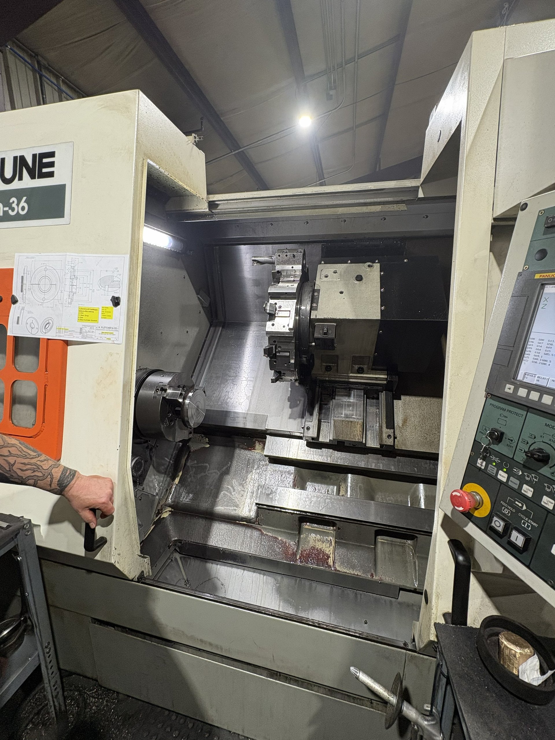 Industrial CNC lathe machine with cream and dark gray exterior, control panel visible on right side, workshop setting with overhead lighting