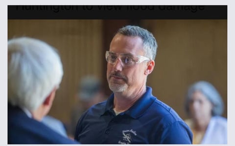 Man with glasses wearing a blue polo shirt speaking to another person at an indoor event
